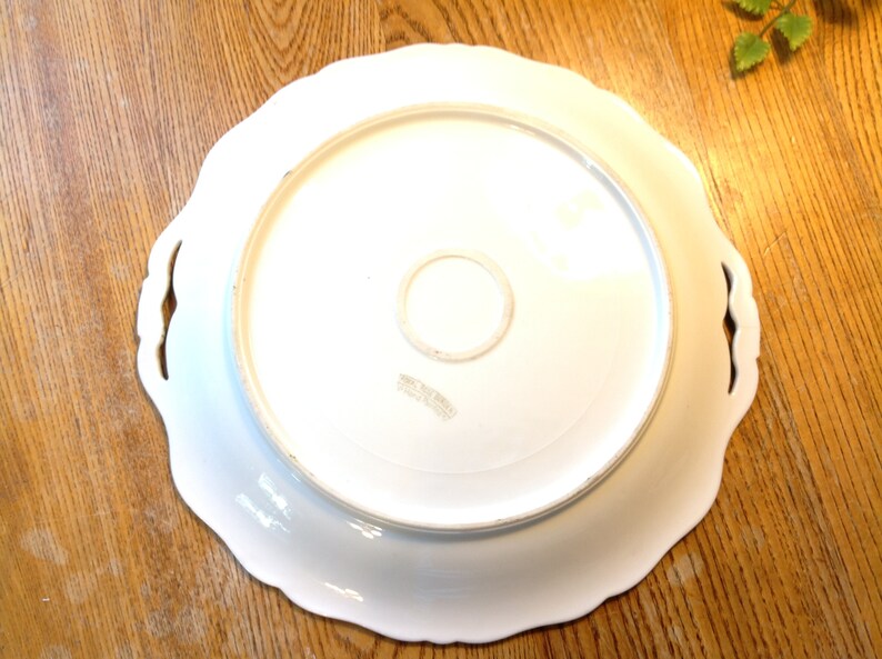 Puede incluir: Plato de servir de cer&aacute;mica blanca con bordes festoneados y dos asas. La parte inferior del plato tiene una marca circular con el texto "Made in Italy".