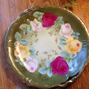 Puede incluir: Un plato de porcelana verde y dorado con un dise&ntilde;o floral decorativo con rosas rosas, amarillas y blancas. El plato tiene un borde festoneado y un borde dorado.