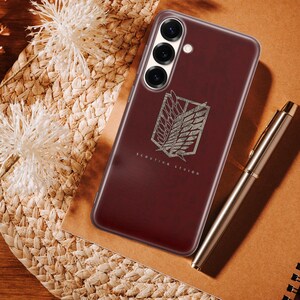 Może przedstawiać: Bordowe etui na telefon ze srebrnym emblematem i napisem "Scouting Legion". Etui leży na tkanej powierzchni z długopisem i notatnikiem. Telefon ma trzy obiektywy.