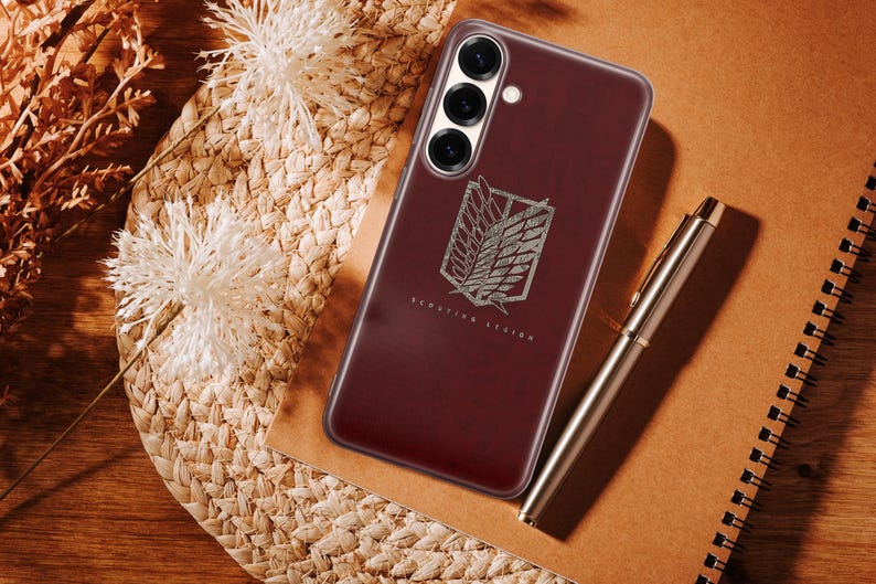 Może przedstawiać: Bordowe etui na telefon ze srebrnym emblematem i napisem "Scouting Legion". Etui leży na tkanej powierzchni z długopisem i notatnikiem. Telefon ma trzy obiektywy.