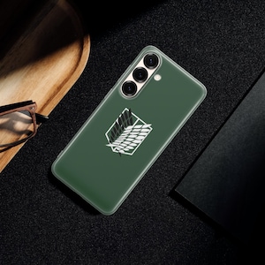 Może przedstawiać: Zielone etui na telefon z białym emblematem, prawdopodobnie na smartfona. Etui leży na ciemnej powierzchni, obok drewnianej tacy i czarnego notatnika. Emblemat to stylizowany wz&oacute;r.