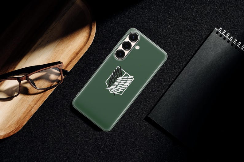 Może przedstawiać: Zielone etui na telefon z białym emblematem, prawdopodobnie na smartfona. Etui leży na ciemnej powierzchni, obok drewnianej tacy i czarnego notatnika. Emblemat to stylizowany wz&oacute;r.