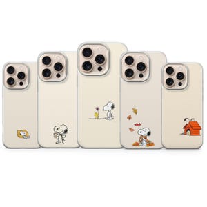 May include: A collection of five beige iPhone cases featuring Snoopy and Woodstock illustrations. Each case showcases a different scene, including Snoopy with coffee, in autumn leaves, and near his doghouse. The cases have clear edges.