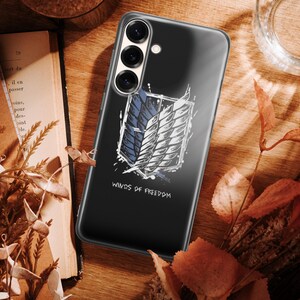 Może przedstawiać: Czarne etui na telefon z białym i niebieskim emblematem skrzydeł, z napisem "Winds of Freedom" poniżej. Leży na drewnianej powierzchni z suchymi liśćmi i książką.