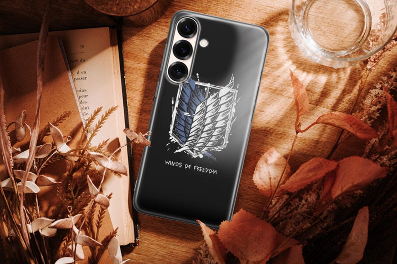 Może przedstawiać: Czarne etui na telefon z białym i niebieskim emblematem skrzydeł, z napisem "Winds of Freedom" poniżej. Leży na drewnianej powierzchni z suchymi liśćmi i książką.