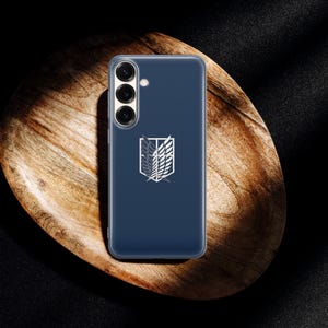 Może przedstawiać: Granatowe etui na telefon z białym emblematem, na drewnianej tacy. Emblemat przedstawia stylizowany wz&oacute;r skrzydeł i herbu. Etui ma przezroczystą osłonę ochronną.