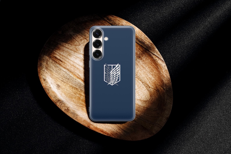 Może przedstawiać: Granatowe etui na telefon z białym emblematem, na drewnianej tacy. Emblemat przedstawia stylizowany wz&oacute;r skrzydeł i herbu. Etui ma przezroczystą osłonę ochronną.