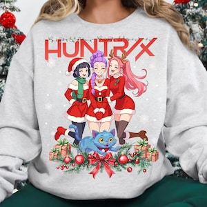 May include: A light gray sweatshirt featuring a festive anime-style illustration. The design includes three figures in red Santa outfits, a blue cat, and the word "HUNT/X" in red. The image is decorated with Christmas ornaments and snowflakes.