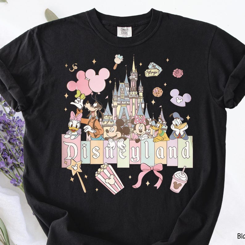 May include: Black t-shirt featuring a colorful Disneyland graphic. The design includes Disney characters like Mickey and Minnie Mouse, a castle, and the word "Disneyland" in pastel colors. Additional elements include popcorn, a bow, and a drink.
