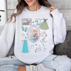 Könnte beinhalten: Hellgraues Sweatshirt mit Illustrationen aus dem Film Frozen. Das Design umfasst Bilder von Elsa und Anna, Kleidern, Schneeflocken und den Texten "Let it go" und "Worth melting for."