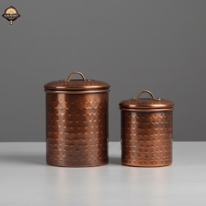 Op de afbeelding: Twee koperkleurige metalen potten met een gehamerde textuur. De potten hebben deksels met kleine, messingkleurige handgrepen. De grotere pot is cilindrisch en de kleinere is korter. De achtergrond is effen grijs.