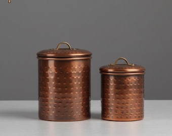 May include: Two copper-colored metal canisters with hammered texture. The canisters have lids with small, brass-colored handles. The larger canister is cylindrical, and the smaller one is shorter. The background is a solid gray.