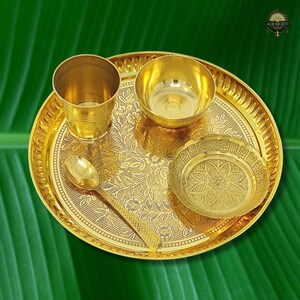 May include: A golden brass puja thali set with a floral design. The set includes a large round tray, a small cup, a bowl, a small plate, and a spoon. The thali is designed for religious ceremonies.