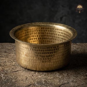 Pode incluir: Uma tigela de latão martelado com uma borda larga e uma superfície texturizada. A tigela tem um tom dourado e assenta sobre uma superfície de pedra. A tigela é provavelmente usada para cozinhar ou servir.