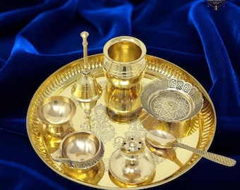 May include: A gold-coloured puja thali set, featuring a round tray and various religious items. The set includes a bell, a small bowl, a spoon, a cup, a small dish, and a lamp. The items are arranged on a gold-coloured tray with a floral design.