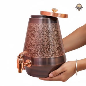 May include: A copper-colored water dispenser with an ornate floral pattern. The dispenser has a spigot, a lid with a crystal-like knob, and is held by two hands. The dispenser is designed with a wide base and tapers upwards.