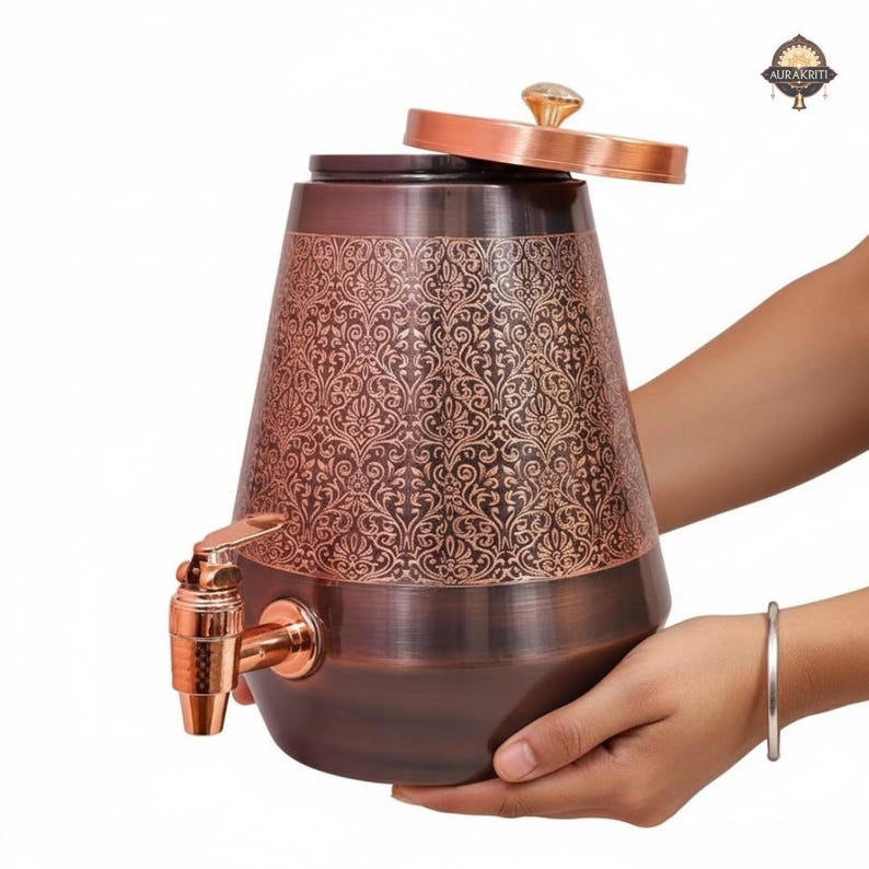 May include: A copper-colored water dispenser with an ornate floral pattern. The dispenser has a spigot, a lid with a crystal-like knob, and is held by two hands. The dispenser is designed with a wide base and tapers upwards.