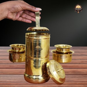 Könnte beinhalten: Ein Kaffee-Filter-Set aus Messing mit zylindrischem Körper, Deckel und kleinen Schalen. Das Set wird auf einer Holzoberfläche präsentiert. Eine Hand hält den Filter offen. Der Markenname "AURAKRITI" ist auf dem Filter sichtbar.