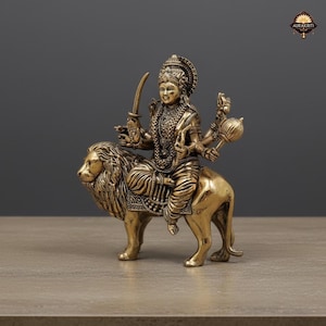 May include: A detailed brass statue of a deity riding a lion. The figure is adorned with intricate jewelry and holds a sword and other symbolic objects. The statue has a golden finish and sits on a neutral surface.