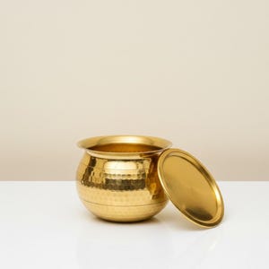 May include: A gold-colored metal pot with a matching lid. The pot has a rounded shape with a hammered texture on the lower half and horizontal bands. The lid is oval and rests beside the pot. The pot is on a white surface.