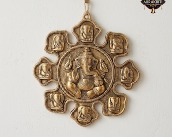 May include: A gold-colored, circular wall hanging featuring a central depiction of the Hindu deity Ganesha, surrounded by eight smaller elephant-headed figures. The intricate design is detailed with relief carvings and a textured finish, and is suspended by a natural fiber cord.