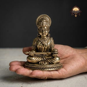 May include: A brass statue of a seated deity, adorned with intricate details, including a halo and ornate jewelry. The figure holds a musical instrument and a small pot. The statue is displayed on a hand, showcasing its size and craftsmanship.