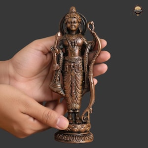 Pode incluir: Uma estátua de cor bronze de uma divindade, segurada nas mãos de uma pessoa. A estátua apresenta detalhes intrincados, incluindo uma auréola, roupas ornamentadas e um arco. A figura está em pé sobre uma base decorativa. É um item religioso ou espiritual.