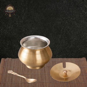 May include: A brass-coloured pot with a silver-coloured rim, a matching lid, and a small spoon. The pot has horizontal etched lines around its body. The lid has a small knob. The spoon has a decorative handle. The items are on a bamboo mat.