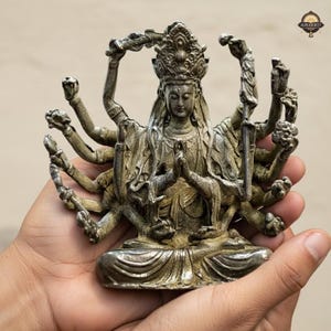Puede incluir: Una detallada estatua de color plateado de una deidad con múltiples brazos, que lleva una corona y ropa ornamentada. La estatua se sostiene en las manos de una persona, mostrando su intrincado diseño y artesanía. La estatua es una representación de una figura religiosa.