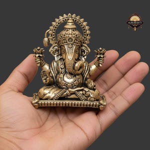 Może przedstawiać: Szczeg&oacute;łowa mosiężna statua hinduskiego b&oacute;stwa Ganesha, siedzącego w medytacyjnej pozie. Statua posiada skomplikowane rzeźbienia, dużą głowę i wiele ramion, trzymających kwiaty lotosu. Figurka trzymana jest w dłoni na ciemnoszarym tle.
