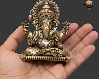 May include: A detailed brass statue of the Hindu deity Ganesha, seated in a meditative pose. The statue features intricate carvings, a large head, and multiple arms, holding lotus flowers. The figure is held in a hand against a dark gray background.