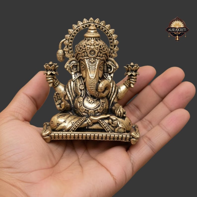 Może przedstawiać: Szczeg&oacute;łowa mosiężna statua hinduskiego b&oacute;stwa Ganesha, siedzącego w medytacyjnej pozie. Statua posiada skomplikowane rzeźbienia, dużą głowę i wiele ramion, trzymających kwiaty lotosu. Figurka trzymana jest w dłoni na ciemnoszarym tle.