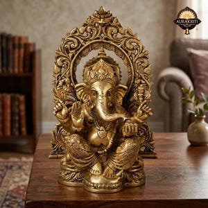 May include: A metallic gold-colored statue of the deity Ganesha, depicted in a seated position with an elephant head and four arms. The figure is framed by an ornate, arched backdrop featuring intricate scrollwork and floral-inspired carvings.