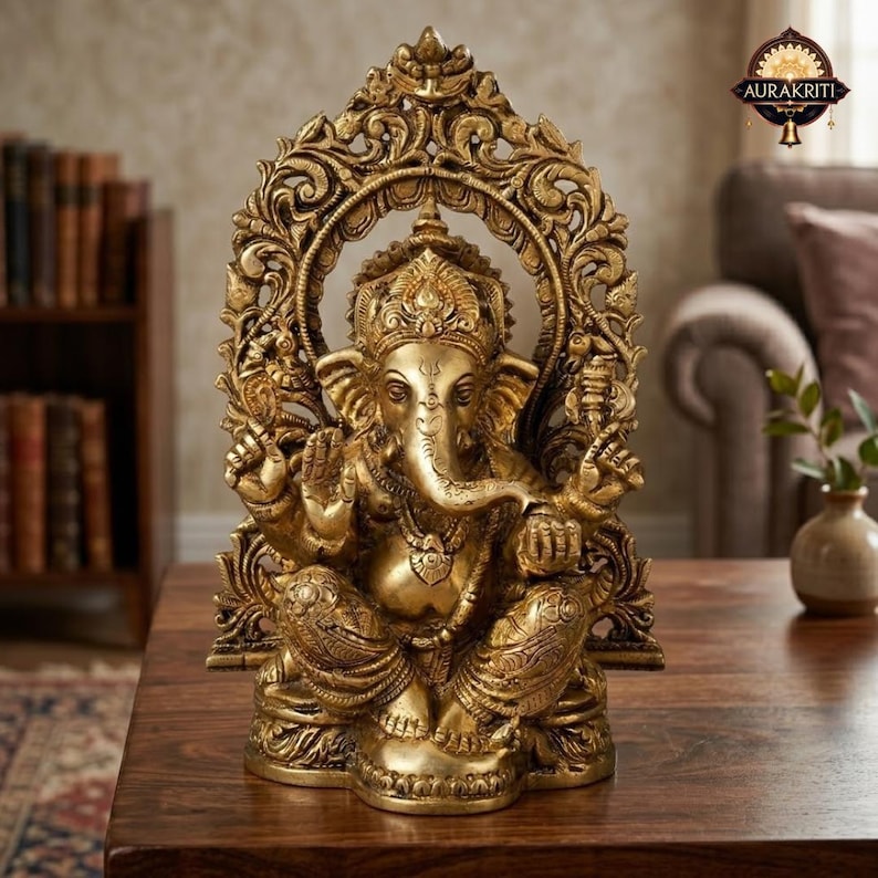 May include: A metallic gold-colored statue of the deity Ganesha, depicted in a seated position with an elephant head and four arms. The figure is framed by an ornate, arched backdrop featuring intricate scrollwork and floral-inspired carvings.