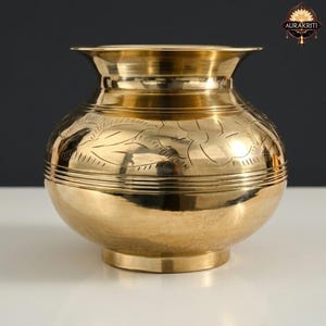 May include: A golden brass pot with a wide, rounded body and a flared rim. The pot features etched floral patterns around the middle and horizontal bands. The pot sits on a small, circular base. The background is a gradient of white to dark gray.