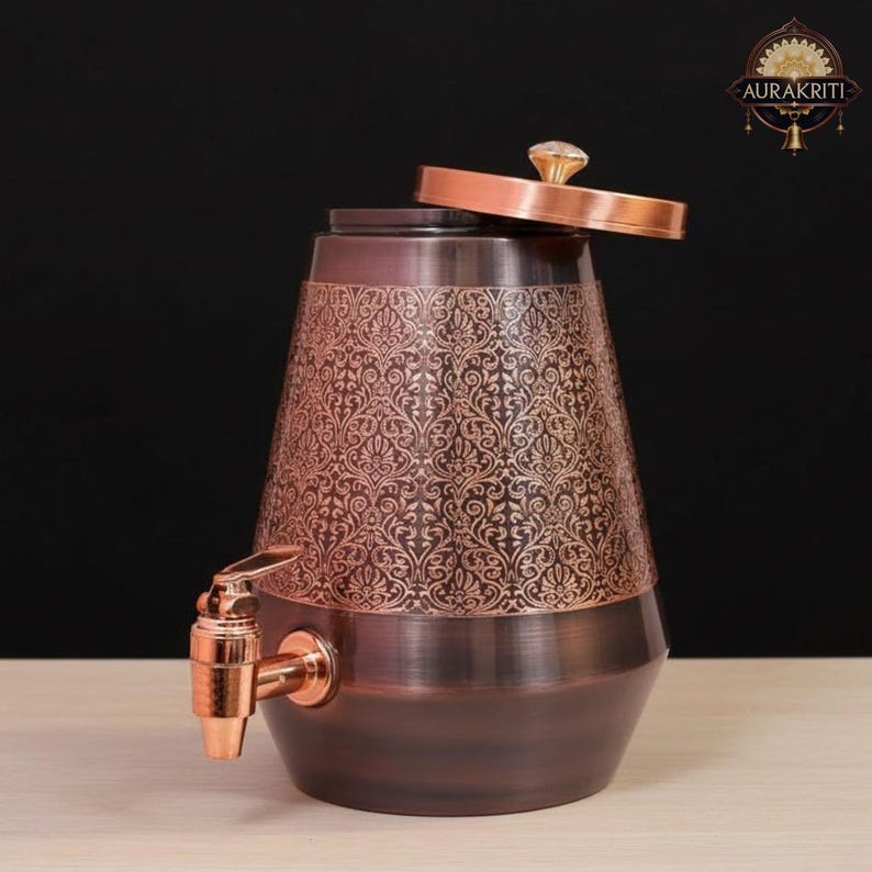 May include: A copper-toned water dispenser with an ornate floral pattern. The dispenser has a spigot and a lid with a crystal-like knob. The design is a mix of dark and light copper tones, creating a vintage aesthetic.