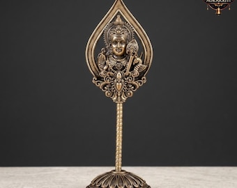May include: A bronze-colored decorative statue of a deity. The statue features a detailed depiction of a figure within a teardrop-shaped frame, mounted on a slender, spiraled stem and a circular base. The Aurakriti logo is visible.