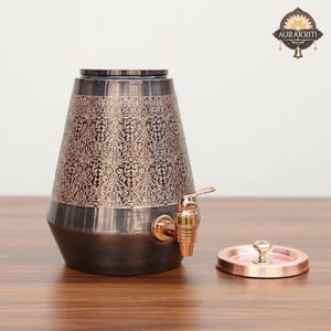 May include: A decorative copper-toned beverage dispenser with an ornate floral pattern. The dispenser has a spigot and a matching lid. The base and top are a darker shade of copper, while the middle section is a lighter copper color.