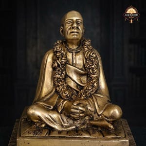 May include: A golden-colored statue of a seated figure, adorned with a floral garland and a necklace. The figure is wearing a robe and is in a meditative pose. The statue is on a square base. The image has the text "AURAKRITI".