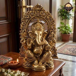 May include: A metallic gold-toned statue of the deity Ganesha seated in a cross-legged position. The figure is framed by an ornate, arched backdrop featuring intricate floral and scrollwork carvings. The statue displays four arms.