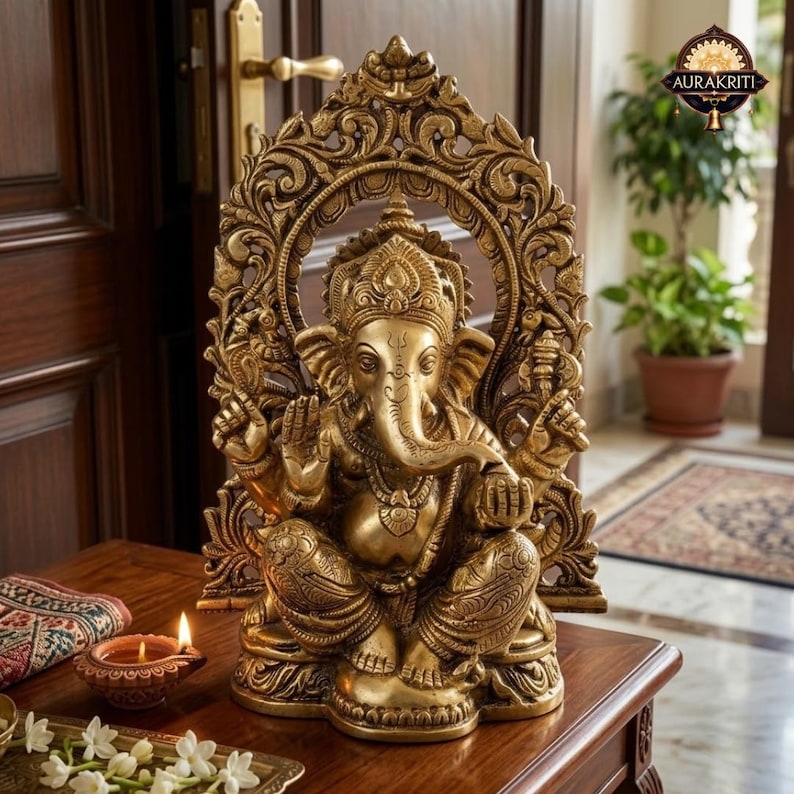May include: A metallic gold-toned statue of the deity Ganesha seated in a cross-legged position. The figure is framed by an ornate, arched backdrop featuring intricate floral and scrollwork carvings. The statue displays four arms.