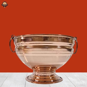 May include: A copper-colored metal bowl with two handles and a pedestal base. The bowl has a wide, open top and a rounded body with horizontal ridges. The background is a solid orange-red color, and the bowl sits on a white wooden surface.