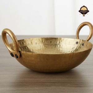Op de afbeelding: Een gehamerde messing kookpot met twee afgeronde handvatten. De pot heeft een brede, ondiepe komvorm en een getextureerd oppervlak. De handvatten zijn met klinknagels bevestigd. De pot is ontworpen om te koken en te serveren.