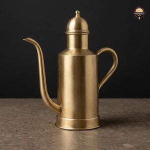 May include: A brushed brass oil dispenser featuring a cylindrical body, a tall domed lid, a curved elongated spout, and a side handle. The metal surface has a matte gold finish. The logo AURAKRITI is visible in the upper right corner of the image.
