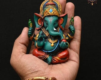 May include: A teal-colored figurine of the Hindu deity Ganesha, adorned with gold and red accents. The statue features intricate details, including a crown, jewelry, and a trunk. The figurine is held in a hand against a dark background.