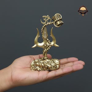 May include: A brass-coloured religious statue depicting a trident, the Om symbol, and a beaded chain with a drum-like object. The statue is on a textured base, held in a hand against a grey background.