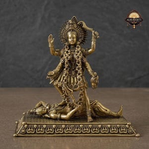 Puede incluir: Una estatua de latón de una deidad de múltiples brazos, sosteniendo una espada y una cabeza cortada, de pie sobre una figura que yace sobre una base decorativa. La estatua tiene detalles intrincados y está adornada con guirnaldas y un halo. El logotipo "AURAKRITI" es visible.