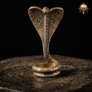 Może przedstawiać: Mosiężna statua kobry z otwartą kapturem, szczegółowymi łuskami i zwiniętą podstawą. Posąg stoi na teksturowanej kamiennej powierzchni na ciemnym tle. Kaptur kobry ma dekoracyjne wiry.