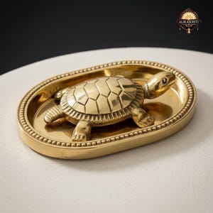 May include: A gold-coloured metal turtle figurine sits in an oval-shaped dish. The turtle has a detailed shell and is surrounded by a beaded border. The dish is on a white surface.