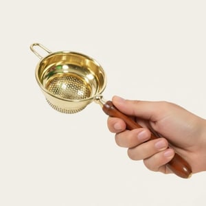 May include: A gold-colored tea strainer with a wooden handle. The strainer has a small handle on the side and a perforated bowl for filtering tea leaves. The wooden handle is a dark brown color.